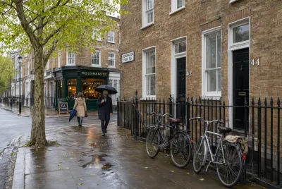 A man with an umbrella and a woman walking on a wet London sidewalk past a brick building and parked bicycles.