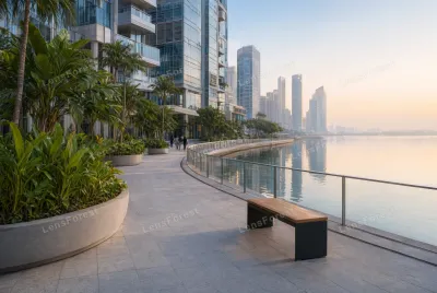 A paved waterfront walkway with a wooden bench, tropical plants in concrete planters, and glass skyscrapers reflecting in the water at sunrise.