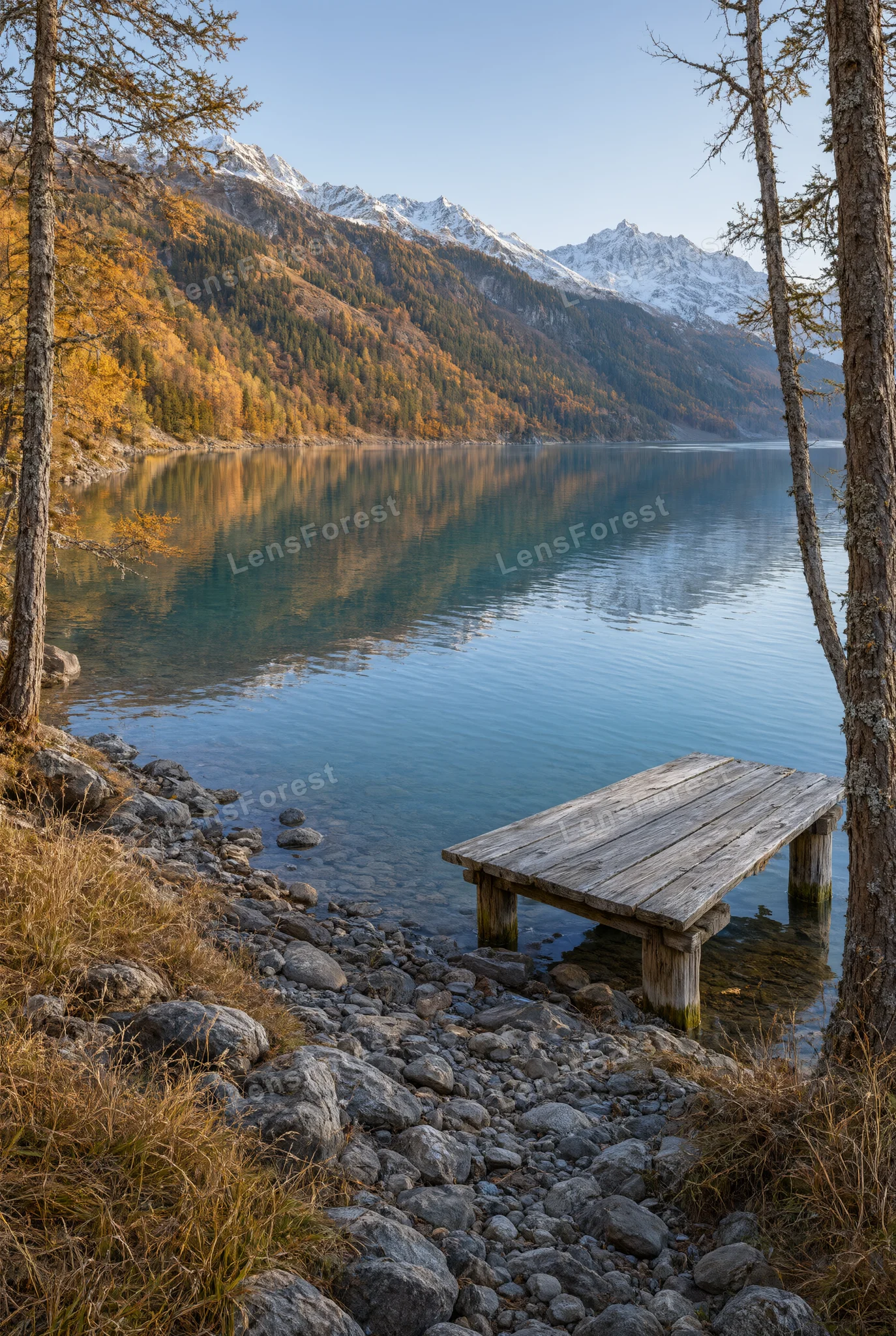 A wooden dock on a rocky shoreline of a calm mountain lake with snow-covered peaks in the distance.