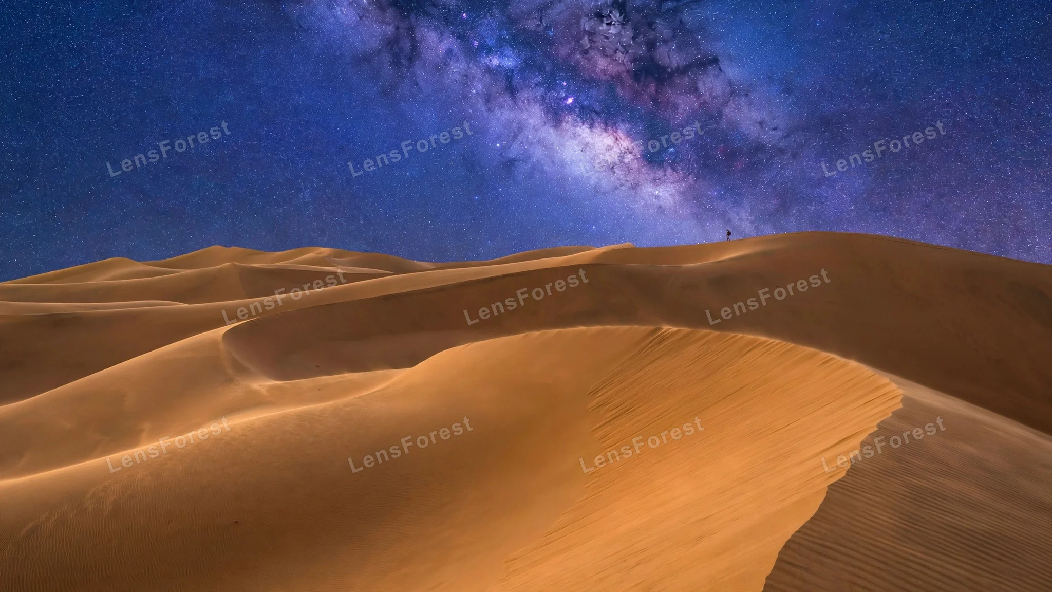 Sweeping golden desert dunes under a brilliant Milky Way star sky with one tiny traveler silhouette on a distant dune ridge
